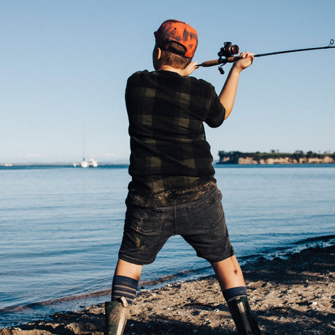 Child wearing Lamington Children's Tide Knee High Socks with boots while standing on shore casting fishing line.