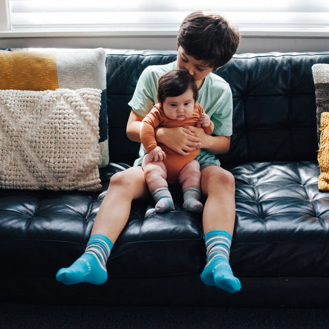 Child wearing Lamington Children's Sky Crew Socks while sitting on couch holding baby.