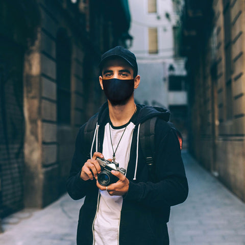 Lifestyle image of model standing in laneway holding a vintage-look camera and wearing a Black Classic MASKiT Face Mask.