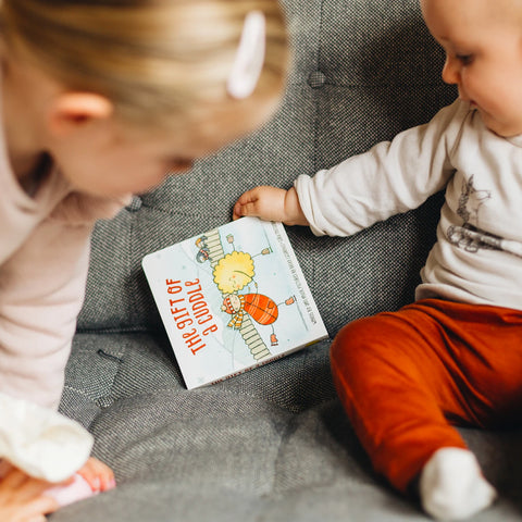 Lifestyle image of a baby and child on a couch holding The Kiss Co.'s The Gift of a Cuddle Board Book.
